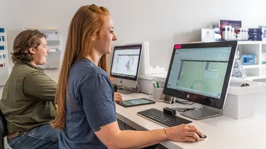 Man and woman sitting at a desk looking at schedules on computers. 