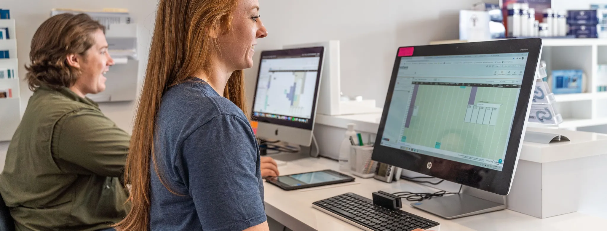 Man and woman sitting at a desk looking at schedules on computers.